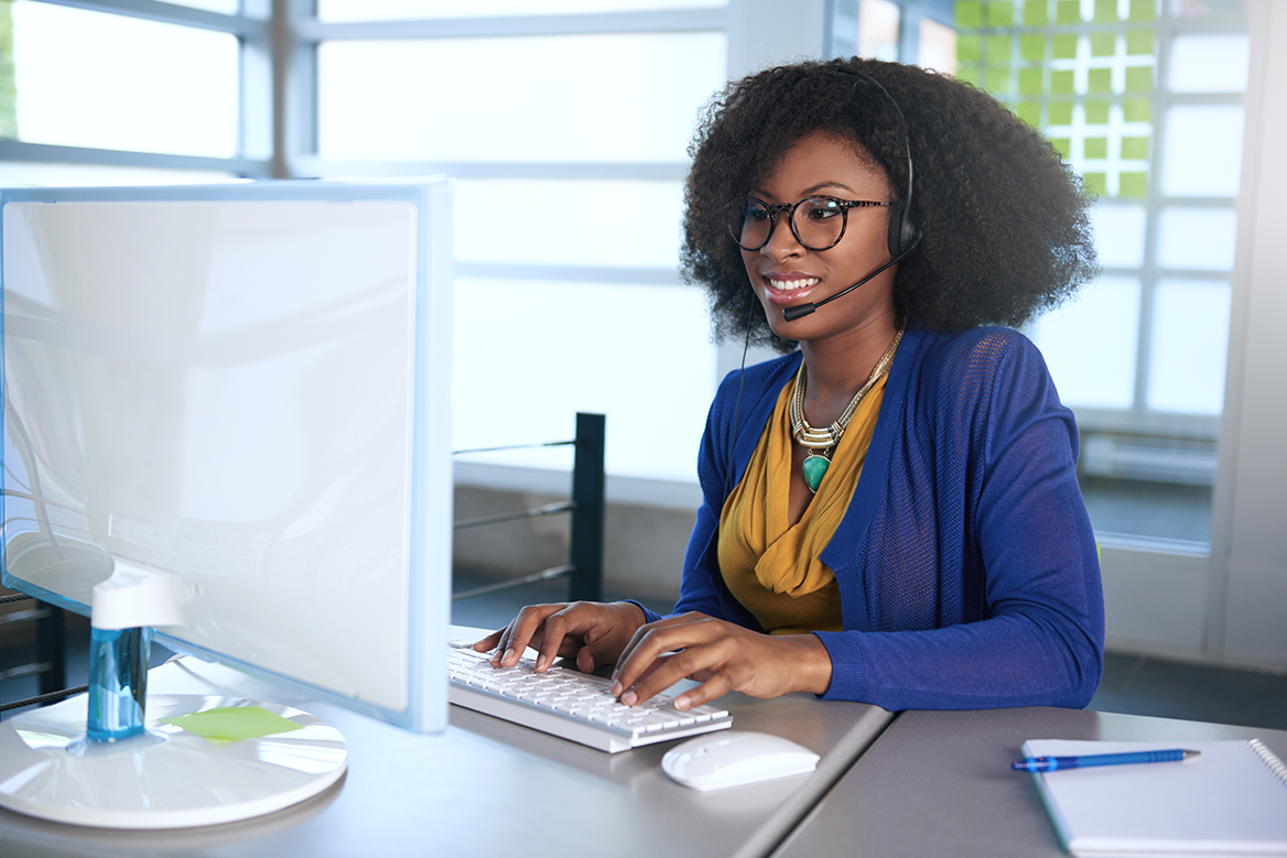 A female phone operator with a headset on a call while typing notes into her computer.