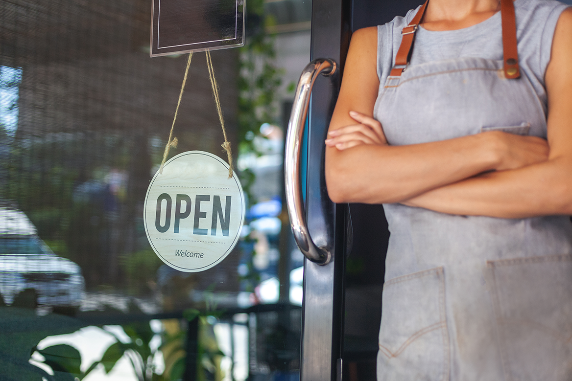 A business owner with arms folded standing next to a door with an open sign.